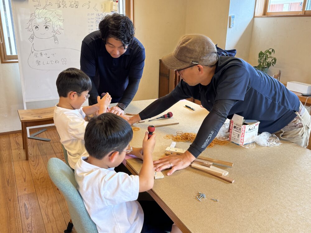 ひだまりフェスのワークショップで、大工さんが子どもたちにゴム鉄砲づくりを優しく教えている様子
